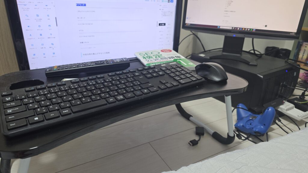 作業環境を写真に撮ったもの。床に置いた小さなテーブル上にワイヤレスキーボード、ワイヤレスマウス。奥には古いテレビとやや小さなモニターによるデュアルモニター、横倒しに置いてあるPCが写っている。作業画面にはブログの執筆画面とChatGPTとの戦略会議の画面。SEO検定1級の公式問題集がテーブルの上に置いてある。 写真の下部には寝床兼作業場になっているマットレスがチラッと写っている。
