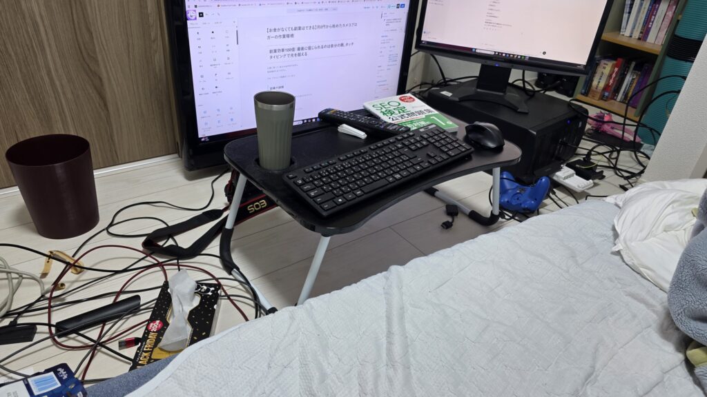 作業環境を写真に撮ったもの。床に置いた小さなテーブル上にワイヤレスキーボード、ワイヤレスマウス。奥には古いテレビとやや小さなモニターによるデュアルモニター、横倒しに置いてあるPCが写っている。作業画面にはブログの執筆画面とChatGPTとの戦略会議の画面。SEO検定1級の公式問題集がテーブルの上に置いてある。 写真の下部には寝床兼作業場になっているマットレスが写っている。ケーブルも床でごちゃごちゃ絡まっている。